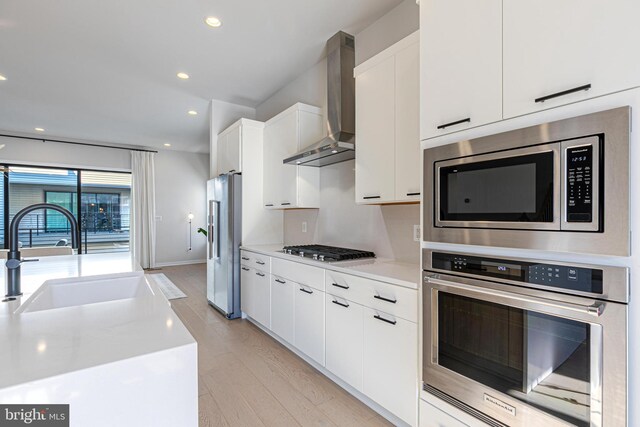 kitchen with appliances with stainless steel finishes, wall chimney exhaust hood, sink, white cabinetry, and light hardwood / wood-style floors