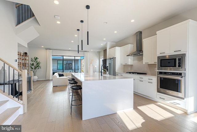 kitchen featuring decorative light fixtures, white cabinets, a kitchen island with sink, wall chimney range hood, and stainless steel appliances