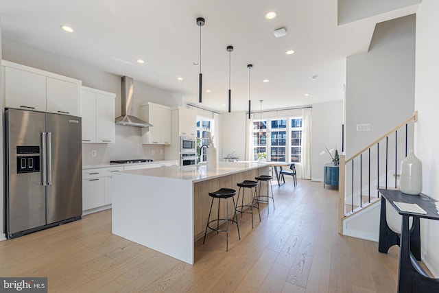 kitchen with pendant lighting, appliances with stainless steel finishes, white cabinets, wall chimney range hood, and a center island with sink