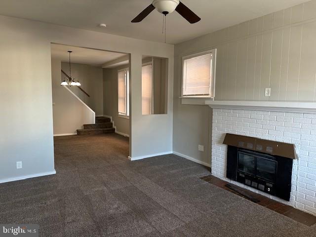 unfurnished living room with dark colored carpet, ceiling fan with notable chandelier, and a brick fireplace