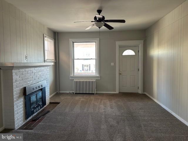 unfurnished living room with radiator, ceiling fan, dark carpet, and a brick fireplace