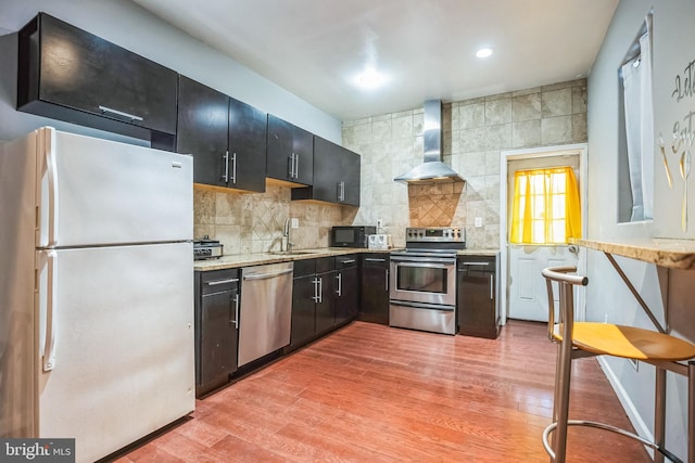 kitchen featuring decorative backsplash, wall chimney exhaust hood, stainless steel appliances, sink, and light hardwood / wood-style flooring