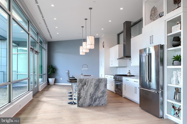 kitchen featuring white cabinetry, wall chimney range hood, hanging light fixtures, a kitchen island with sink, and high quality appliances