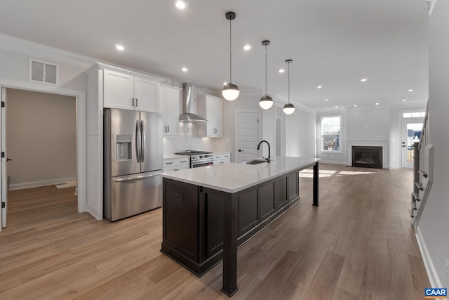 kitchen with sink, hanging light fixtures, wall chimney range hood, a kitchen island with sink, and appliances with stainless steel finishes