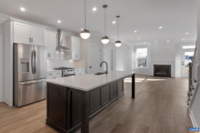 kitchen with wall chimney exhaust hood, stainless steel appliances, sink, hanging light fixtures, and an island with sink