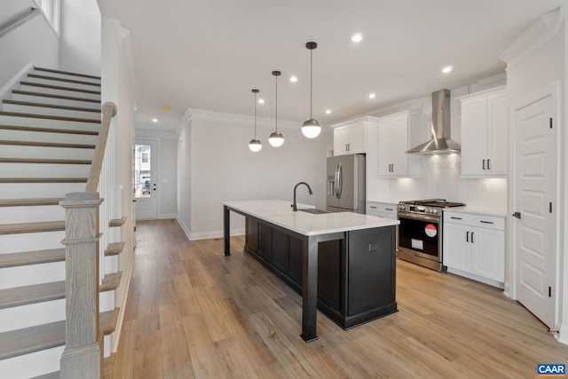 kitchen featuring wall chimney exhaust hood, stainless steel appliances, decorative light fixtures, a center island with sink, and white cabinets