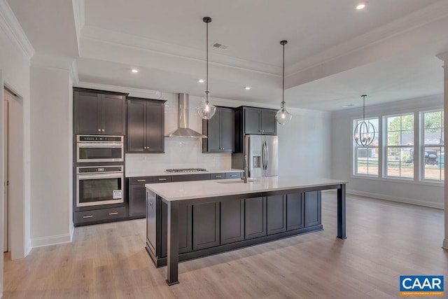 kitchen with pendant lighting, wall chimney range hood, an island with sink, a tray ceiling, and stainless steel appliances