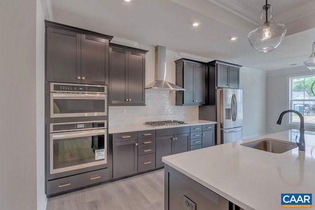 kitchen with stainless steel appliances, crown molding, wall chimney range hood, sink, and pendant lighting