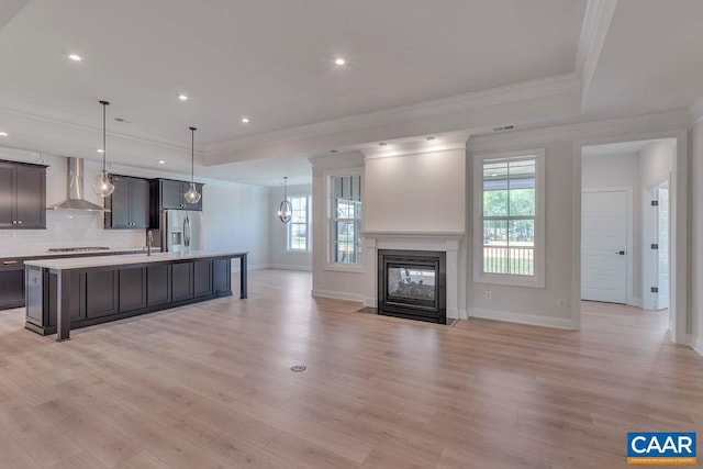 kitchen featuring a kitchen island with sink, stainless steel refrigerator with ice dispenser, wall chimney exhaust hood, light wood-type flooring, and decorative light fixtures