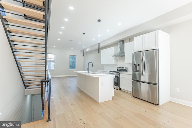 kitchen with white cabinetry, stainless steel appliances, wall chimney range hood, an island with sink, and decorative light fixtures