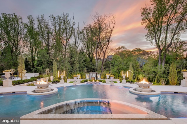 pool at dusk featuring an in ground hot tub, pool water feature, a fire pit, and a patio area