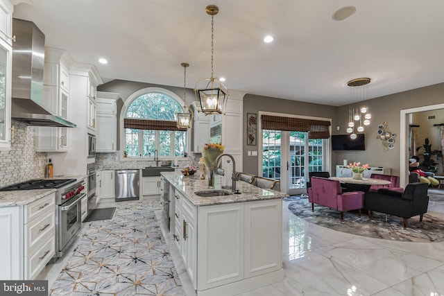 kitchen featuring sink, wall chimney exhaust hood, stainless steel appliances, an island with sink, and white cabinets