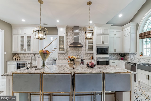 kitchen with appliances with stainless steel finishes, a breakfast bar, wall chimney range hood, hanging light fixtures, and an island with sink