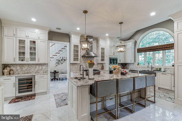 kitchen with a center island with sink, hanging light fixtures, sink, wall chimney exhaust hood, and beverage cooler