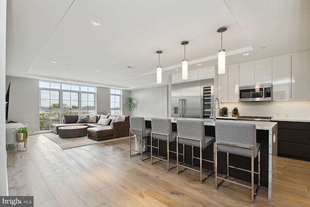 kitchen with a raised ceiling, an island with sink, decorative light fixtures, white cabinetry, and stainless steel appliances