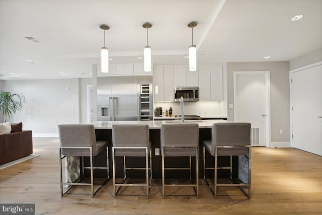 kitchen featuring an island with sink, appliances with stainless steel finishes, decorative light fixtures, light hardwood / wood-style floors, and white cabinetry