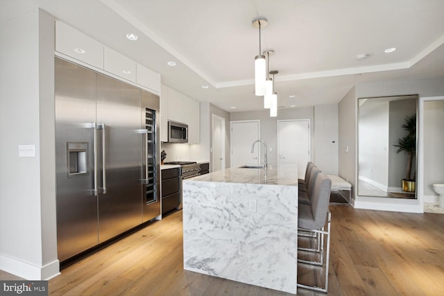 kitchen with a kitchen island with sink, sink, high quality appliances, white cabinetry, and hanging light fixtures