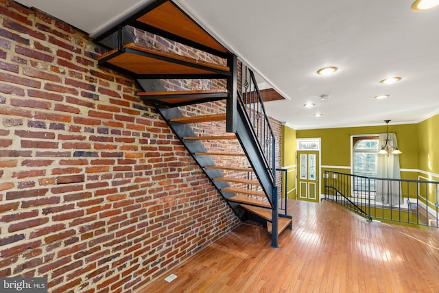 staircase with crown molding, wood-type flooring, brick wall, and a notable chandelier