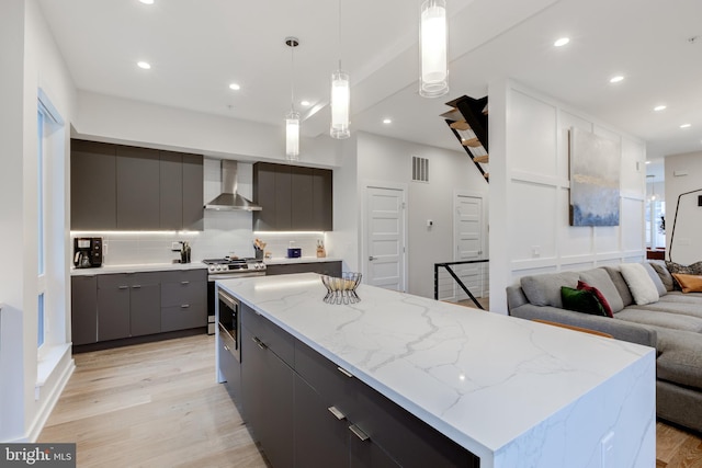 kitchen featuring wall chimney range hood, a kitchen island, light hardwood / wood-style flooring, decorative light fixtures, and stainless steel stove