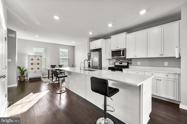 kitchen featuring appliances with stainless steel finishes, white cabinets, a kitchen bar, an island with sink, and dark wood-type flooring