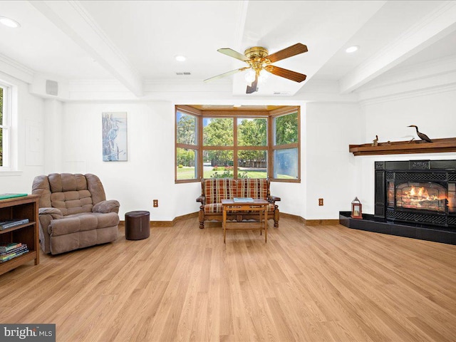 living area with a tile fireplace, ceiling fan, beamed ceiling, crown molding, and light hardwood / wood-style floors