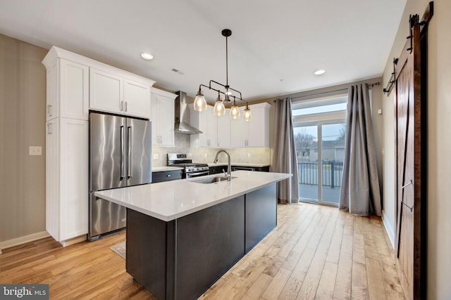 kitchen with pendant lighting, white cabinets, wall chimney exhaust hood, stainless steel appliances, and a kitchen island with sink