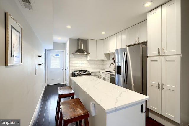 kitchen featuring light stone countertops, wall chimney exhaust hood, stainless steel appliances, a kitchen island, and white cabinets