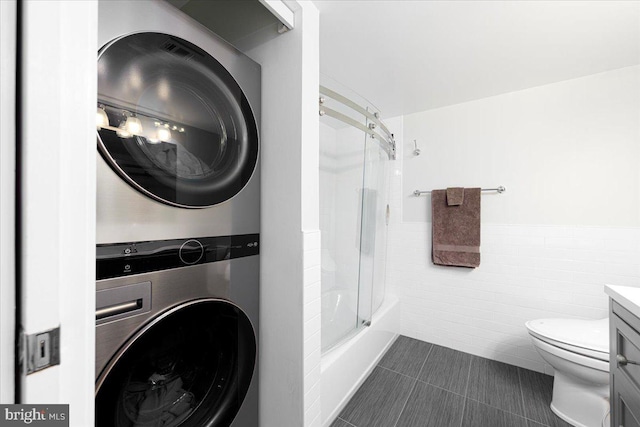 clothes washing area featuring dark tile patterned floors, stacked washing maching and dryer, and tile walls