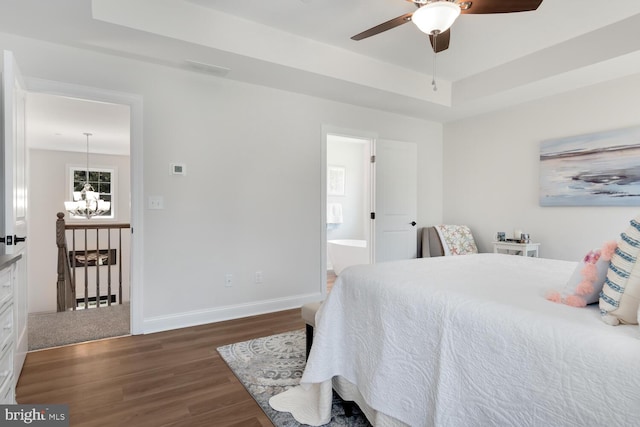 bedroom featuring dark hardwood / wood-style flooring, ensuite bath, ceiling fan with notable chandelier, and a raised ceiling