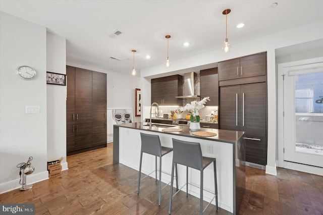 kitchen with dark brown cabinetry, sink, wall chimney exhaust hood, hanging light fixtures, and a kitchen island with sink