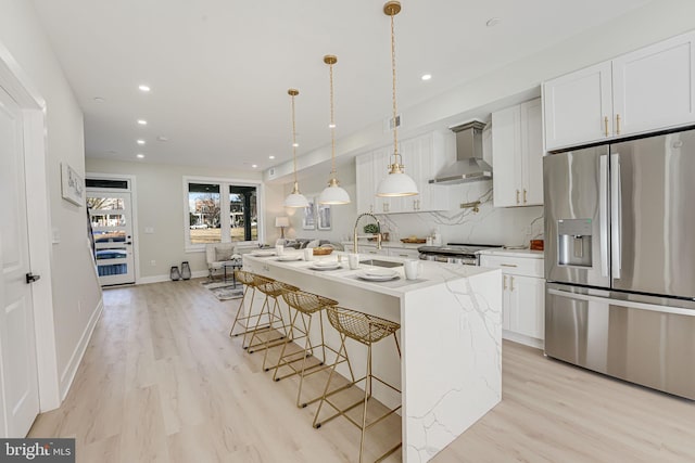 kitchen with light stone countertops, wall chimney exhaust hood, stainless steel appliances, a center island with sink, and white cabinetry