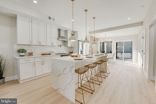 kitchen with white cabinetry, an island with sink, hanging light fixtures, and wall chimney range hood