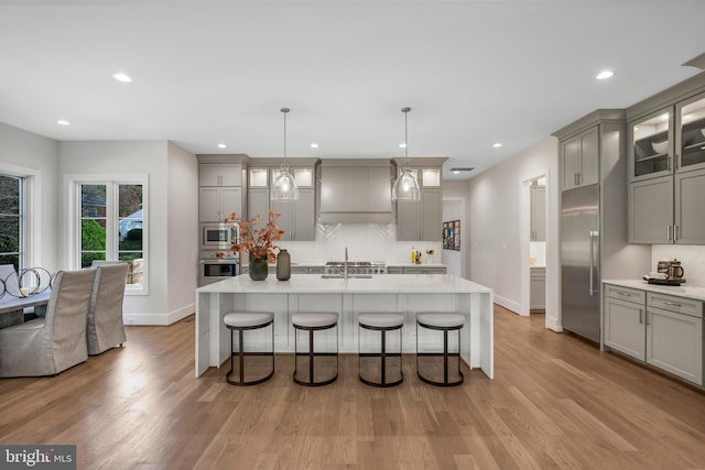 kitchen featuring built in appliances, decorative light fixtures, gray cabinetry, and an island with sink