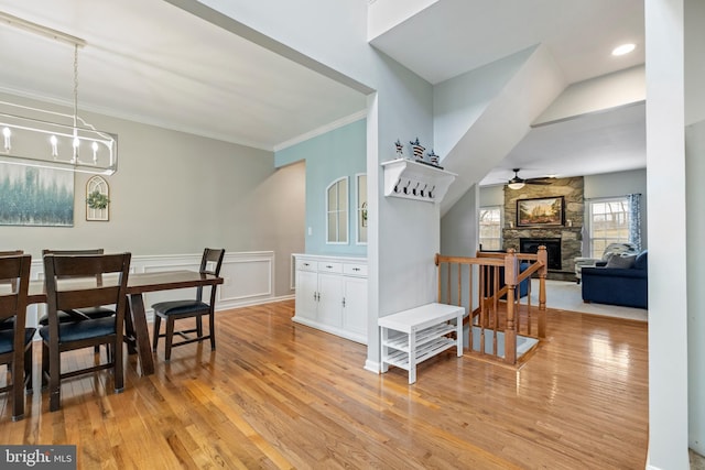 dining space featuring a stone fireplace, ceiling fan with notable chandelier, ornamental molding, and light wood-type flooring