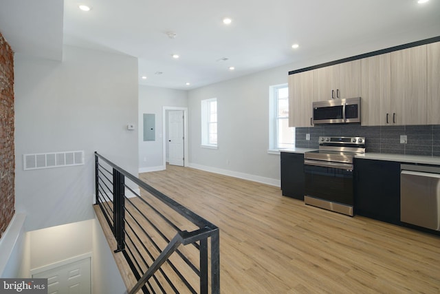 kitchen featuring light brown cabinetry, light wood-type flooring, tasteful backsplash, stainless steel appliances, and electric panel