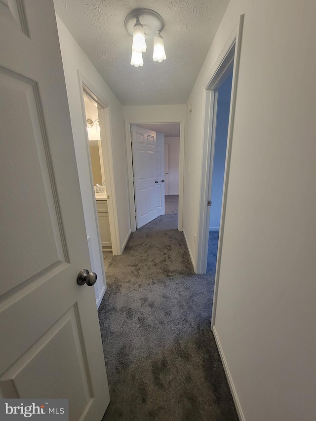 hallway featuring a textured ceiling, dark colored carpet, and baseboards