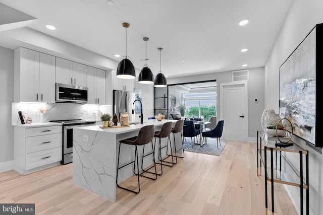 kitchen with appliances with stainless steel finishes, hanging light fixtures, white cabinets, light hardwood / wood-style flooring, and a center island with sink