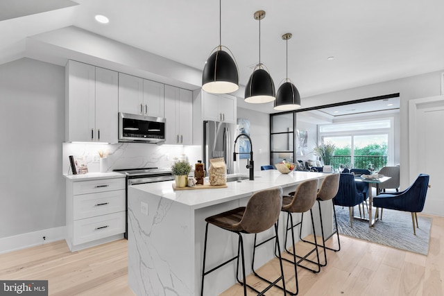 kitchen featuring appliances with stainless steel finishes, an island with sink, decorative backsplash, light hardwood / wood-style floors, and hanging light fixtures