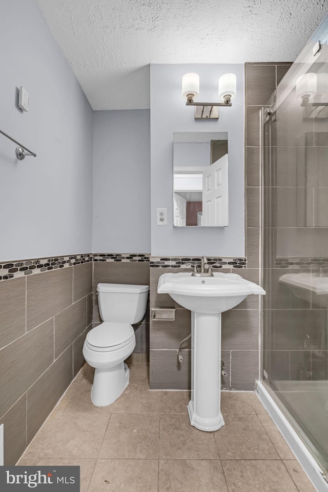 bathroom featuring toilet, a shower with shower door, tile walls, a textured ceiling, and tile patterned flooring