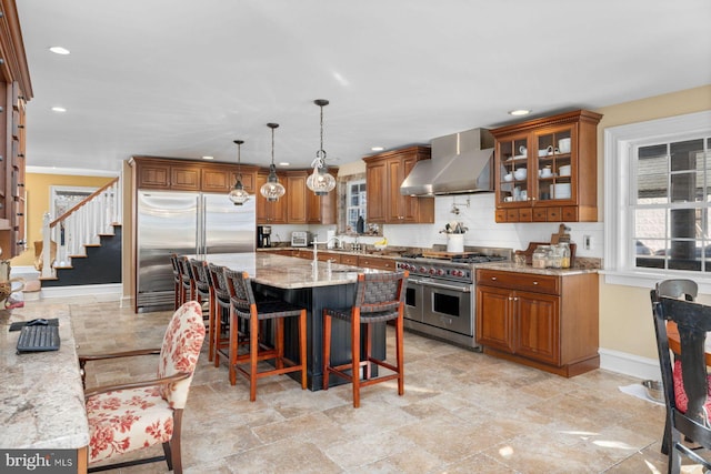 kitchen featuring wall chimney range hood, hanging light fixtures, high end appliances, light stone counters, and a kitchen bar