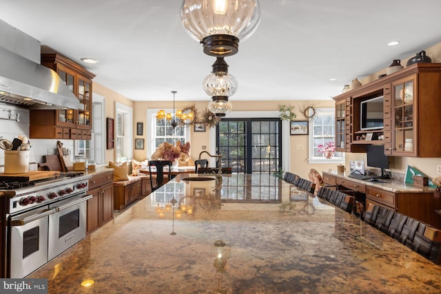 kitchen with hanging light fixtures, range hood, built in desk, light stone countertops, and range with two ovens