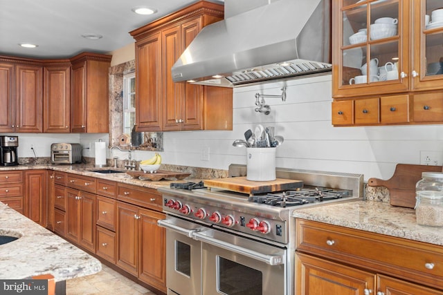 kitchen with light stone counters, wall chimney range hood, sink, and range with two ovens