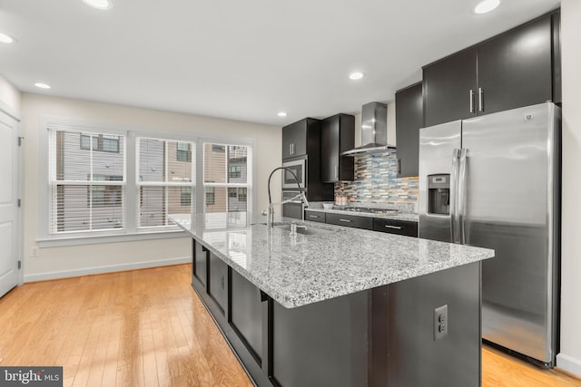 kitchen with appliances with stainless steel finishes, an island with sink, tasteful backsplash, and wall chimney range hood