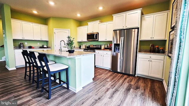 kitchen featuring light stone counters, stainless steel appliances, dark wood-type flooring, a center island with sink, and white cabinetry