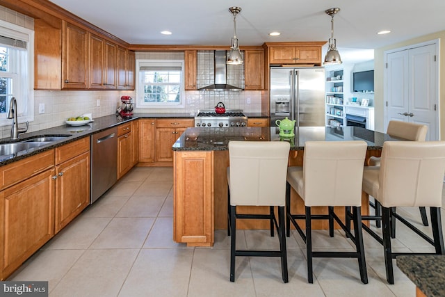 kitchen with wall chimney range hood, sink, appliances with stainless steel finishes, decorative light fixtures, and a kitchen island