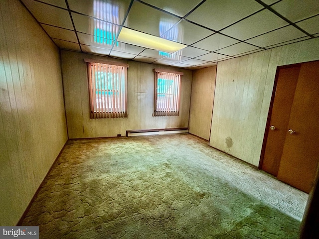 empty room featuring light colored carpet, a drop ceiling, baseboard heating, and wood walls