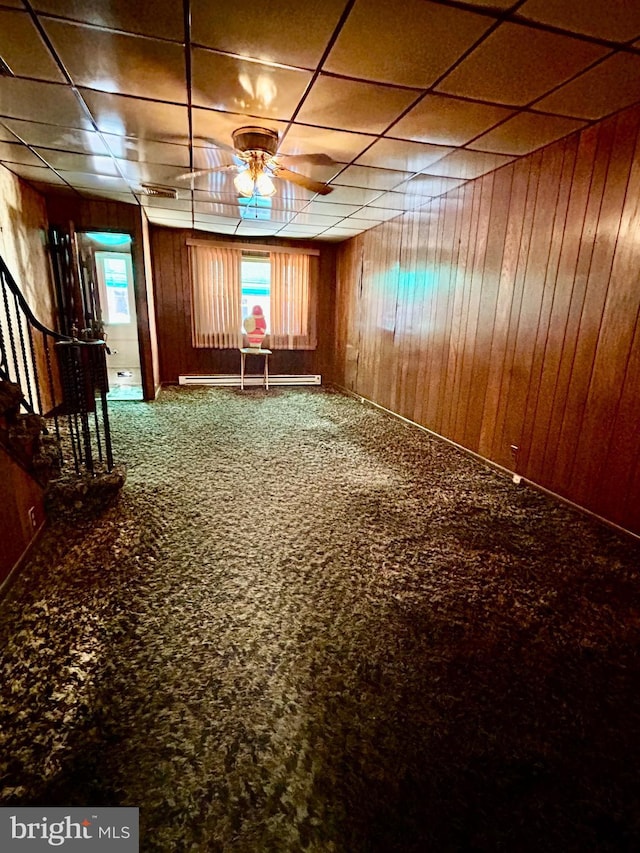 unfurnished room with wood walls, carpet, a baseboard heating unit, ceiling fan, and a drop ceiling