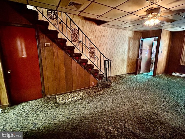 interior space with a paneled ceiling, wooden walls, carpet floors, a baseboard heating unit, and ceiling fan