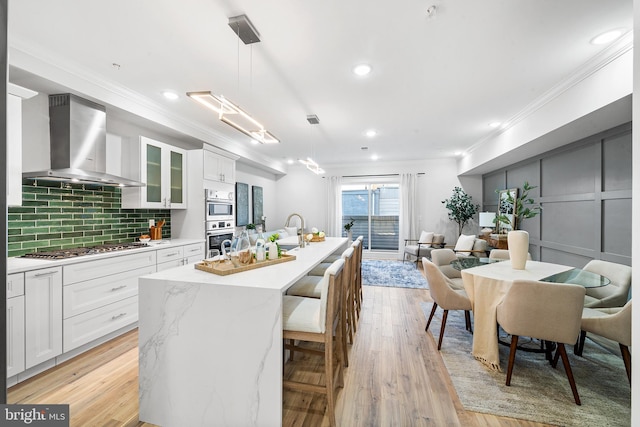 kitchen with a center island with sink, wall chimney range hood, decorative light fixtures, white cabinetry, and stainless steel gas cooktop