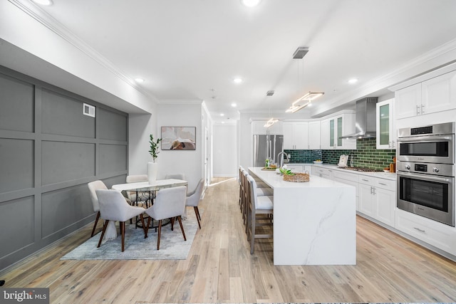 kitchen with wall chimney range hood, decorative light fixtures, a center island with sink, white cabinets, and appliances with stainless steel finishes